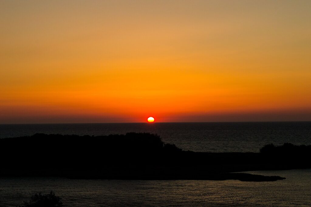 Sonnenaufgang über einem flachen Feld, die Scheibe steht knapp über dem Horizont im Dunst
